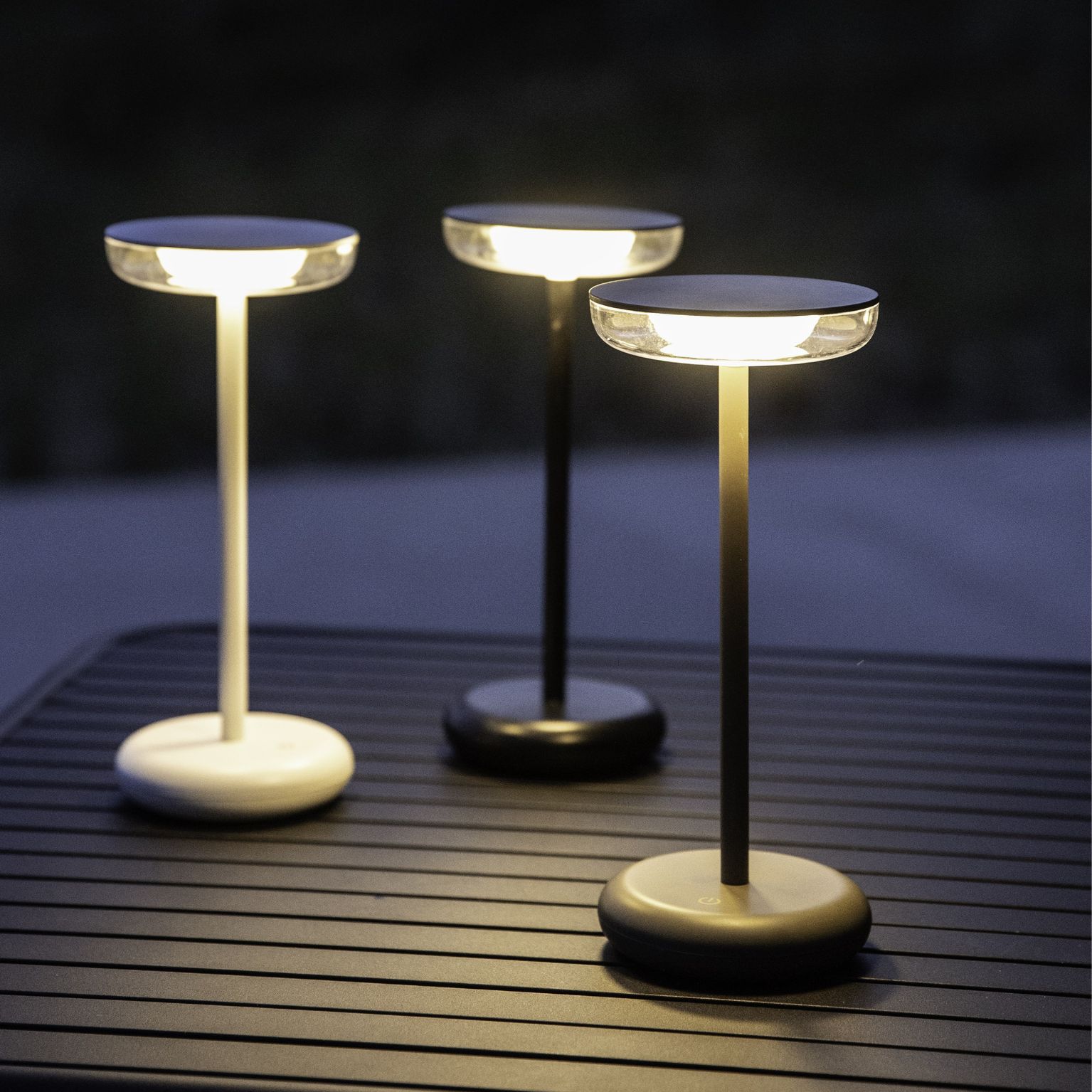 Tre moderna, trådlösa bordslampor med slank design, placerade på ett mörkt utomhusbord med lamellstruktur.