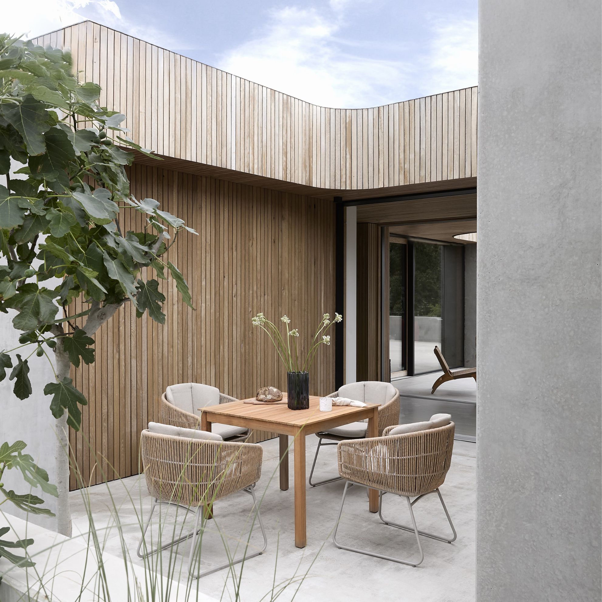 Inblick i en hörna med trädgårdsbord i trä och fyra trädgårdsstolar i beige flätning