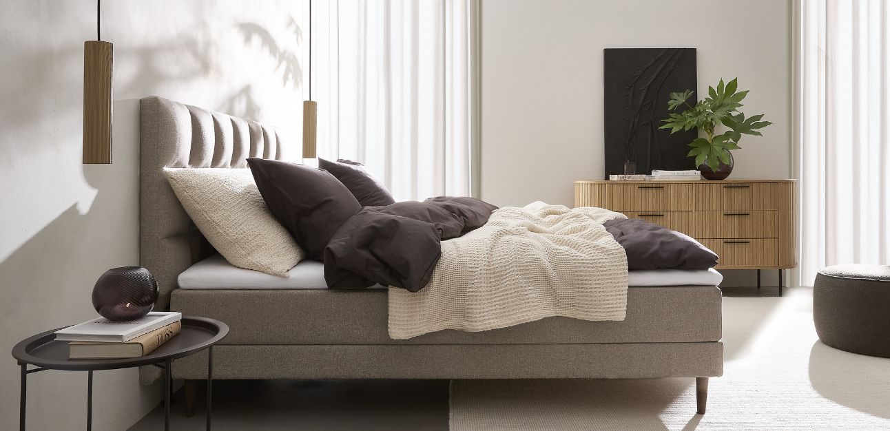 En kontinentalsäng från sidan med sänggavel i snygg design. I sovrummet syns även en träkommod, 2 pendellampor och några andra saker som gör rummet hemtrevligt.