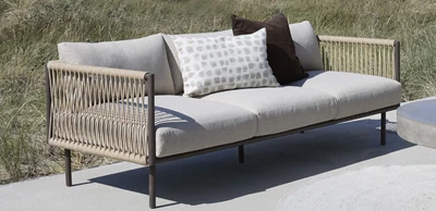 sofa 3 pers.jpg Loungesoffa i flätad design och grå dynor stående på betongunderlag med gräsyta i bakgrunden