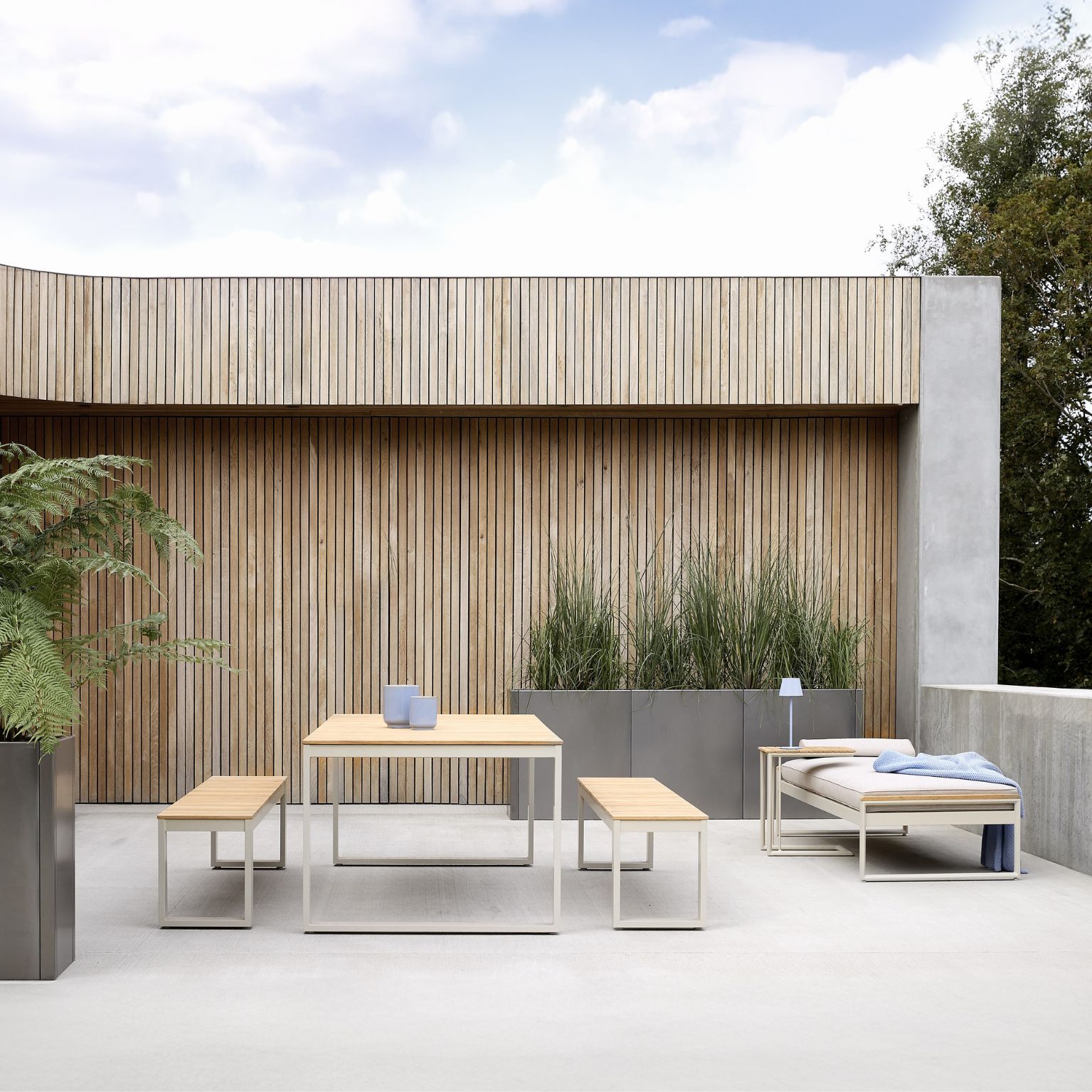 Terrass med vägg i trälameller och betonggolv. Trädgårdsmöblerna består av ett trädgårdsbord, två bänkar samt en solsäng.