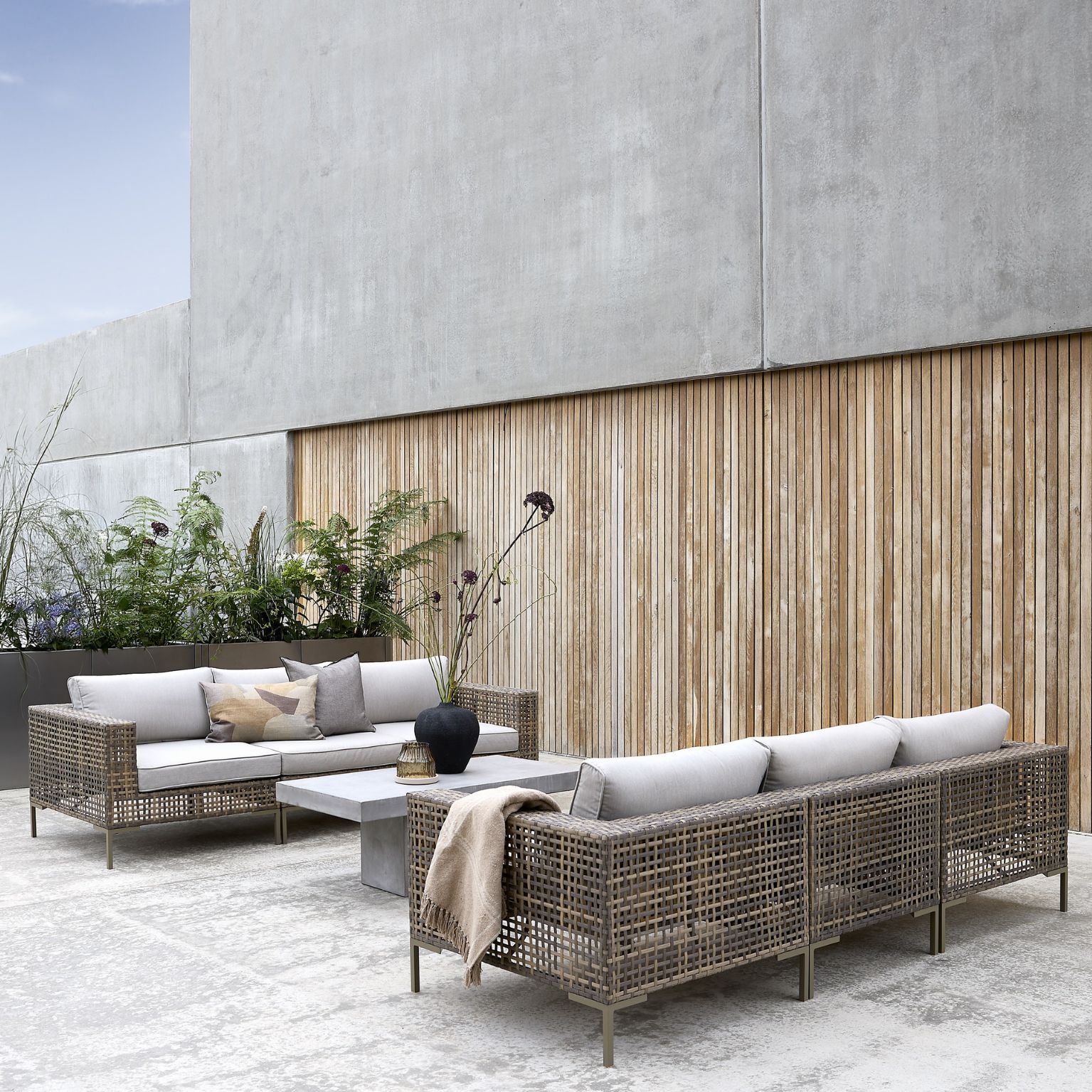 Flätade loungemöbler med grå dynor placerade på betonggolv med trälameller i bakgrunden.