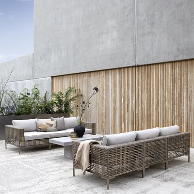 Flätade loungemöbler med grå dynor placerade på betonggolv med trälameller i bakgrunden.