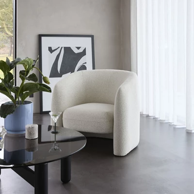 Vit helklädd Loom loungestol i bouclétyg placerad i ett minimalistiskt rum med ett svart glasbord, en grön växt och en konstposter.