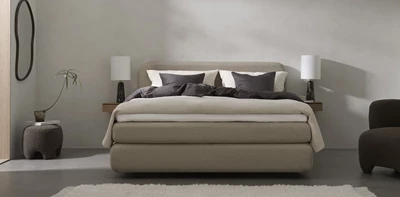 En stilren og minimalistisk soveværelsesindretning med en stor seng i neutrale nuancer. Sengen er redt med mørke og lyse puder samt en kombination af mørkt og lyst sengetøj. Væghængte natborde med sorte bordlamper flankerer sengen. Rummet har en rolig og afdæmpet farvepalette med grå vægge og en lys tæppebelagt gulvflade.