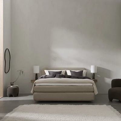En stilren og minimalistisk soveværelsesindretning med en stor seng i neutrale nuancer. Sengen er redt med mørke og lyse puder samt en kombination af mørkt og lyst sengetøj. Væghængte natborde med sorte bordlamper flankerer sengen. Rummet har en rolig og afdæmpet farvepalette med grå vægge og en lys tæppebelagt gulvflade.
