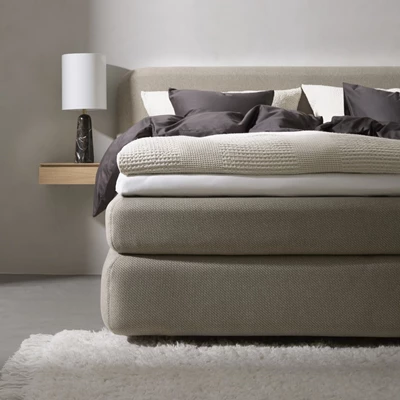 Närbild av en elegant säng med ett beige stoppat huvudgavel. Sängen är bäddad med en kombination av mörkbrunt och offwhite sängkläder, vilket skapar en kontrast.