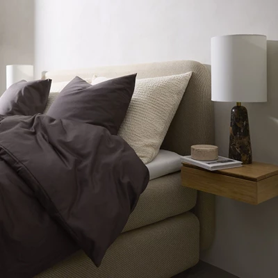 Frontal vy av en lyxig säng med ett mjukt, texturerat beige överdrag. Sängen är uppbyggd av två stoppade madrasser och är bäddad med mörkbruna och ljusa kuddar samt en stickad överkast i en ljus nyans.