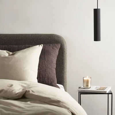 Närbild av en elegant och minimalistisk sänguppsättning med ett mörkbrunt, stoppat huvudgavel. Sängen är bäddad med beige sängkläder och har en kombination av ljusa och mörkbruna kuddar, där den mörka har en quiltad textur.