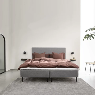 Minimalistiskt sovrum med en grå dubbelsäng, brunt sängkläder och svarta vägglampor. En spegel och ett litet bord med växt dekorerar rummet