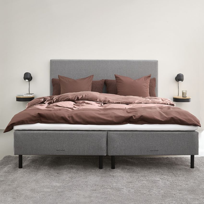 En stilren säng med ljusgrå stoppad sängram och svarta ben. Sängen är bäddad med bruna sängkläder och matchande kuddar