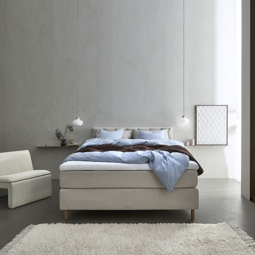 Ett elegant och harmoniskt sovrum med en stor, ljusgrå stoppad kontinentalsäng, bäddad med ljusblå och mörkbruna sängkläder.