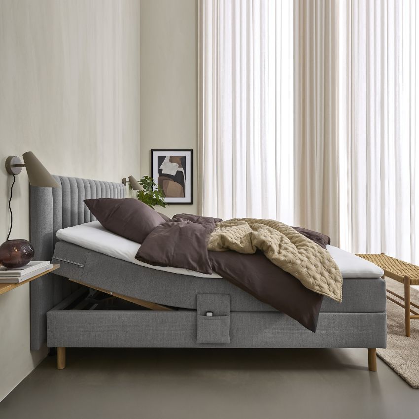 En modern ställbar säng med mörkbrun stoppad sängram. Sängen är bäddad med mörkbruna kuddar och sängkläder samt en quiltad beige pläd som ger extra värme och komfort