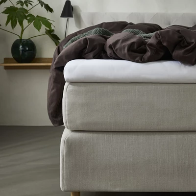 Närbild av en lyxig kontinentalsäng med en ljus, stoppad sängram och träben. Sängen är bäddad med mörkbruna sängkläder och en djupt grön, stickad pläd som tillför textur och värme