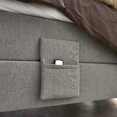 Närbild av en grå ställbar säng med en integrerad tygficka på sidan, där en fjärrkontroll förvaras. Sängen är bäddad med mörkbruna sängkläder och en mjuk, beige quiltad pläd som tillför textur och värme