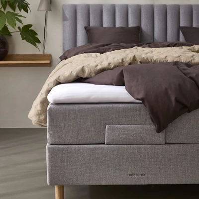 Närbild av en grå ställbar säng med en hög, stoppad sänggavel med vertikala paneler. Sängen är bäddad med mörkbruna sängkläder och en beige quiltad pläd som tillför värme och textur. En praktisk tygficka på sidan av sängen ger plats för förvaring av en fjärrkontroll, vilket understryker sängens funktionalitet.