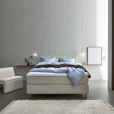 Ett modernt och harmoniskt sovrum med en stor, ljusgrå stoppad kontinentalsäng. Sängen är bäddad med ljusblå och mörkbruna sängkläder samt en stickad pläd som tillför värme och textur