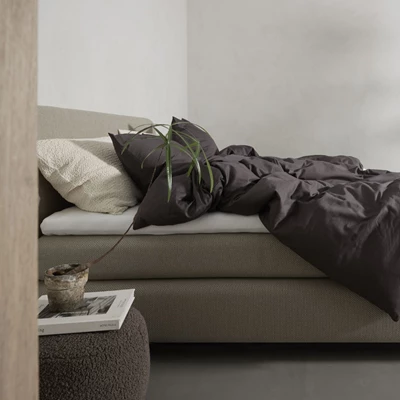 Närbild av en modern säng med en beige stoppad ram och mörkbruna sängkläder som ger ett elegant och diskret uttryck. Kuddarna består av en kombination av mörkbruna och cremefärgade stickade textilier som tillför textur och kontrast