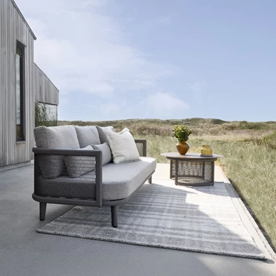 Stilfull terrass med utsikt över sanddyner: Ett bekvämt utomhus loungeområde med en grå och vit matta, modern soffa och dekorativt bord, perfekt för avslappning i naturen.