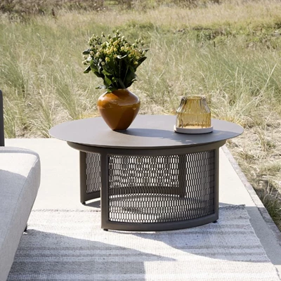 Stilrent utomhusbord, dekorerat med en rustik keramikvas fylld med gröna blommor och en dekorativ glasbehållare, omgiven av en neutral, vävd matta på en terrass med utsikt över vilda gräsmarker.