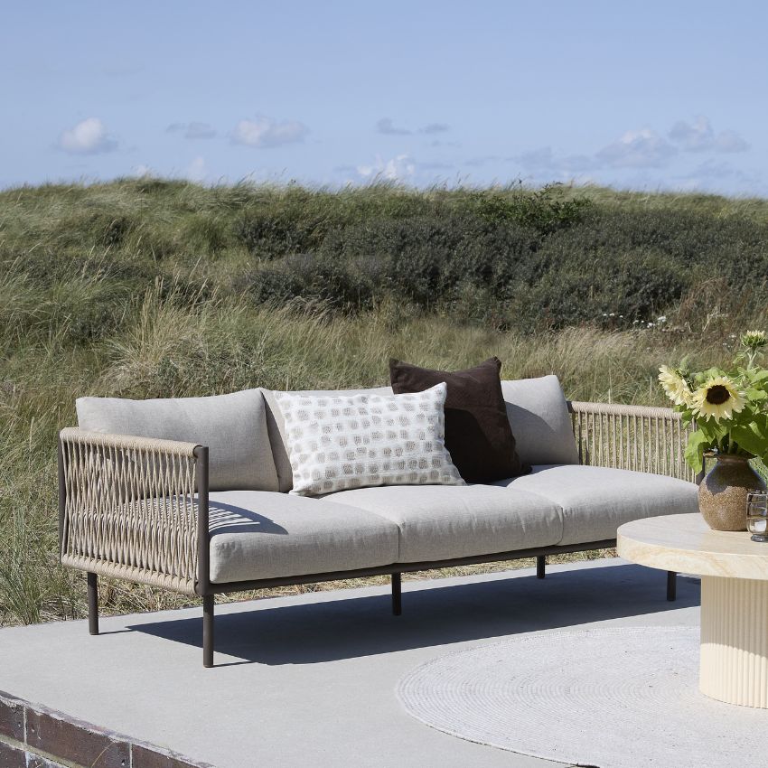 ChatGPT sagde: Utomhussoffa i dansk design med kuddar och sidobord vid grönskande landskap.