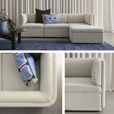 Modern beige soffa med schäslong och dekorativa kuddar på en mörkblå matta framför en vit gardin.