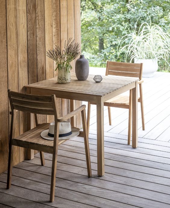 Trädgårdsbord i trä med en vas och stol på en träveranda omgiven av gröna växter