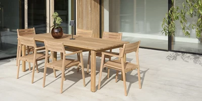 Design uden navn - 2025-02-25T113133.810.jpg Utomhus matområde med stort träbord och sex stolar, beläget på en ljus terrass vid en byggnad med stora fönster