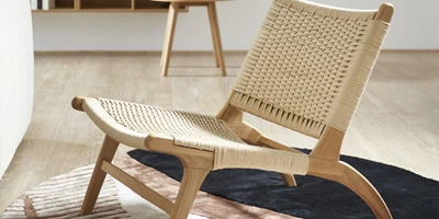 Design uden navn - 2025-02-25T134705.162.jpg Minimalistisk loungestol i ljust trä och flätad sits, placerad på en matta i en stilren interiör.