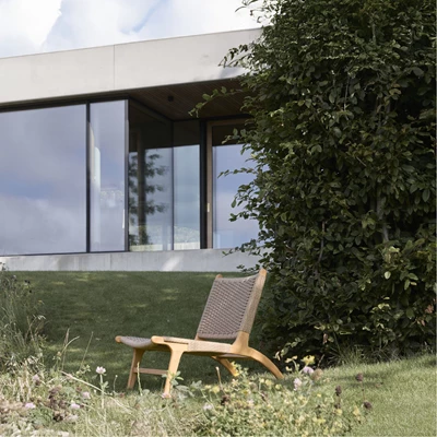 Utomhus loungestol i trä och flätad sits, placerad på en grön gräsmatta framför ett modernt hus med stora glaspartier.
