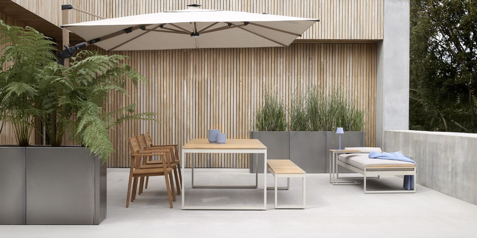 Modern terrass med matbord i trä och metall, en bänk och stolar under ett stort parasoll. Gröna växter i höga krukor fulländar den stilrena inredningen.