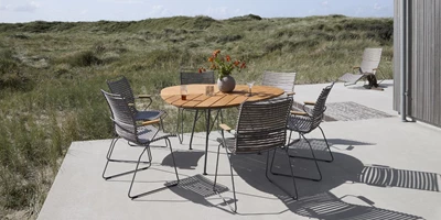 Design uden navn - 2025-03-02T202040.440.jpg Utomhus matplats med svarta flätade stolar runt ett runt träbord, placerat på en terrass med dynbakgrund.