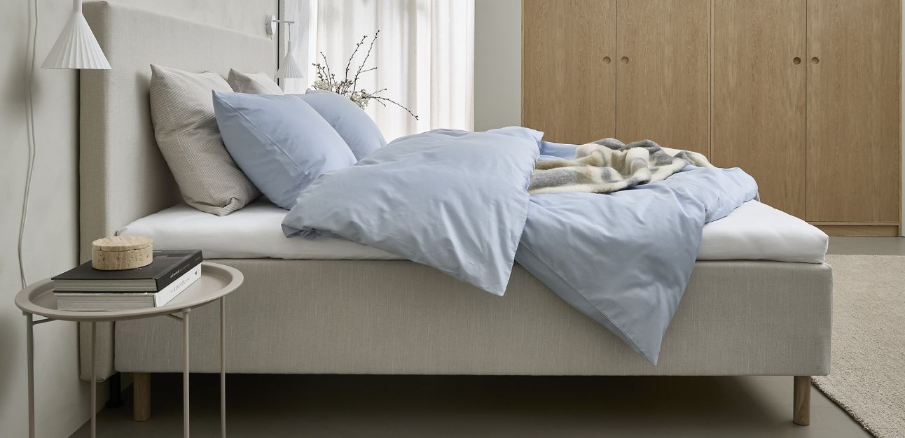 Ljus sovrumsinredning med en beige säng, ljusblått sängkläder och ett nattduksbord i metall med böcker och en lampa.