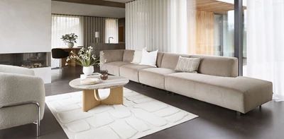 Lyxigt vardagsrum med en lång beige soffa, runda soffbord i trä och sten, omgiven av stora fönster med utsikt över naturen