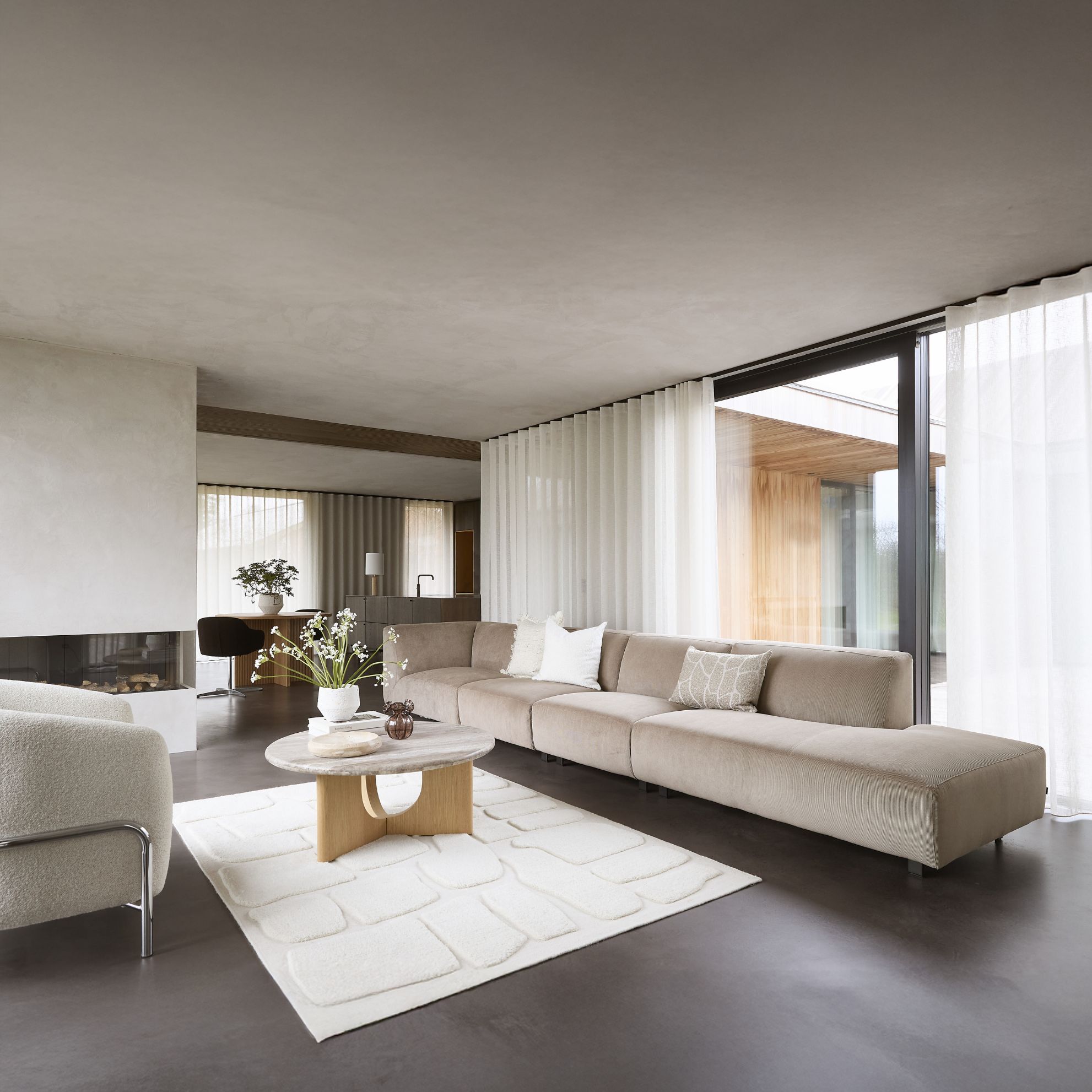 Lyxigt vardagsrum med en lång beige soffa, runda soffbord i trä och sten, omgiven av stora fönster med utsikt över naturen