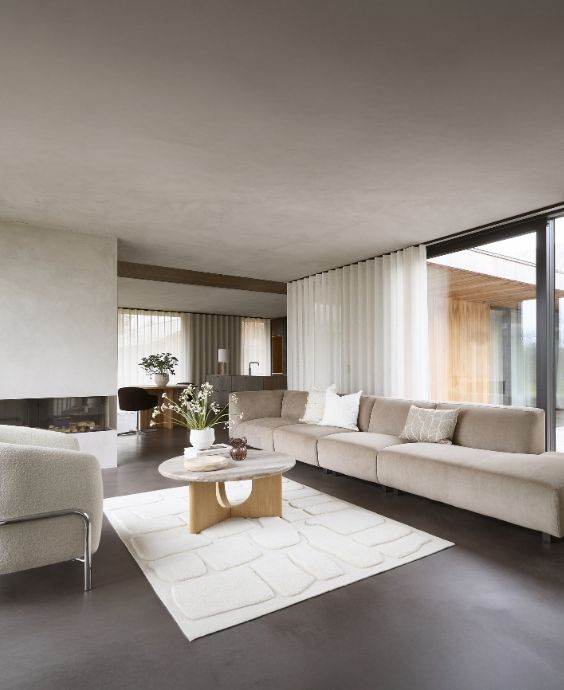 Lyxigt vardagsrum med en lång beige soffa, runda soffbord i trä och sten, omgiven av stora fönster med utsikt över naturen.