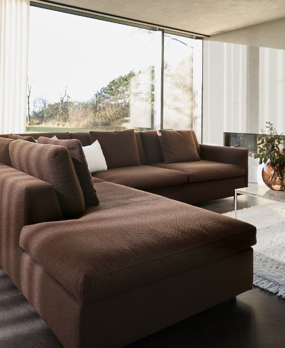 Modernt vardagsrum med mörkbrun hörnsoffa, stor golvlampa och öppna ytor med stora panoramafönster.