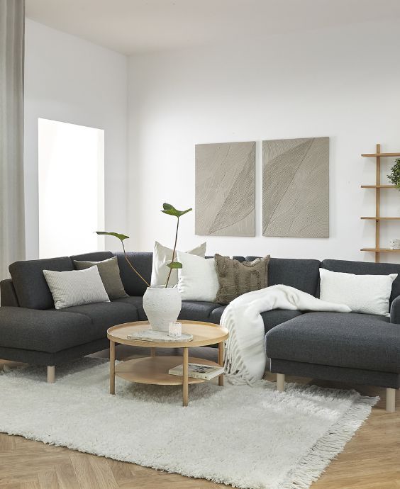 Mysigt vardagsrum med en L-formad mörkgrå soffa, runt träbord i mitten, omgiven av en mjuk vit matta och dekorerad med kuddar. Modern konst hänger på den ljusa väggen.