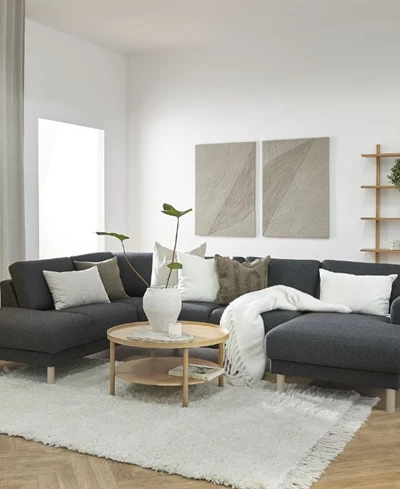 Mysigt vardagsrum med en L-formad mörkgrå soffa, runt träbord i mitten, omgiven av en mjuk vit matta och dekorerad med kuddar. Modern konst hänger på den ljusa väggen.