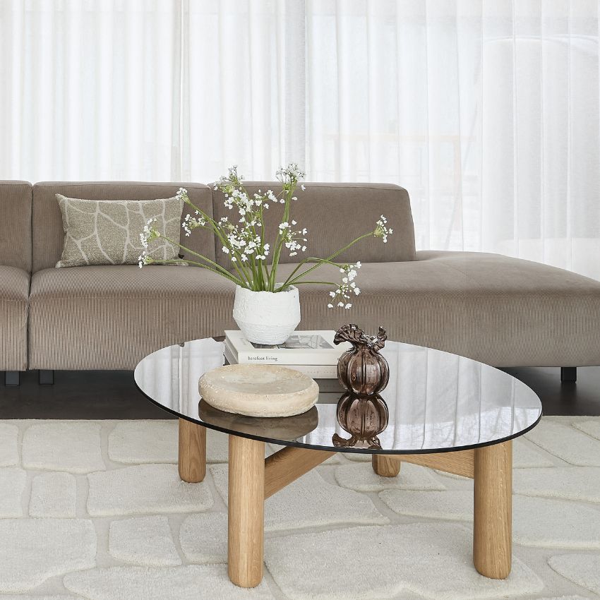 Ett vardagsrum med en mörkgrå soffa och ett runt soffbord i glas framför, där en vas med blommor och dekorativa element står