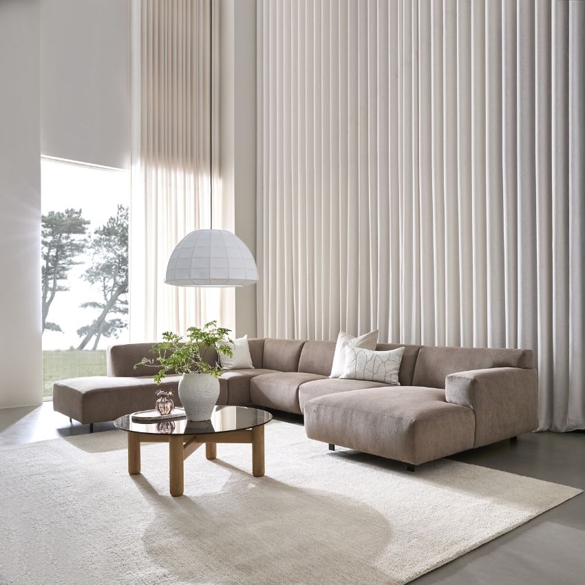 Rymligt vardagsrum med en stor, ljusgrå soffgrupp och ett lågt träbord framför. En vit matta och långa vita gardiner fulländar den ljusa och luftiga atmosfären.