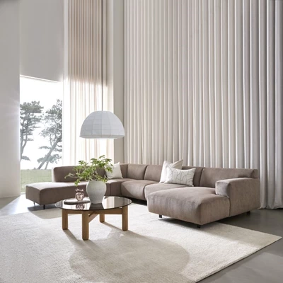 Rymligt vardagsrum med en stor, ljusgrå soffgrupp och ett lågt träbord framför. En vit matta och långa vita gardiner fulländar den ljusa och luftiga atmosfären.