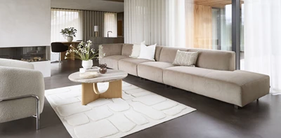 Modernt vardagsrum med en lång, beige modulsoffa vid en eldstad, komplett med ett runt soffbord i trä och marmor.