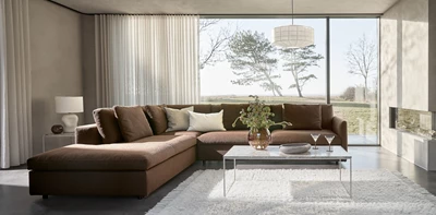 Lyxigt vardagsrum med en stor, mörkbrun modulsoffa och ett soffbord i glas, framför stora fönster med utsikt över naturen.