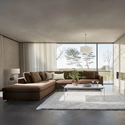 Lyxigt vardagsrum med en stor, mörkbrun modulsoffa och ett soffbord i glas, framför stora fönster med utsikt över naturen.