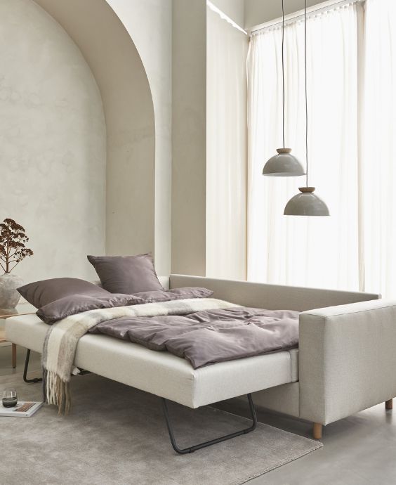 Minimalistiskt sovrum med en stor beige säng under ett böjt fönster med hängande pendellampor och mjukt grått sängkläder. Rummet utstrålar lugn och elegans med sina neutrala färger och mjuka belysning