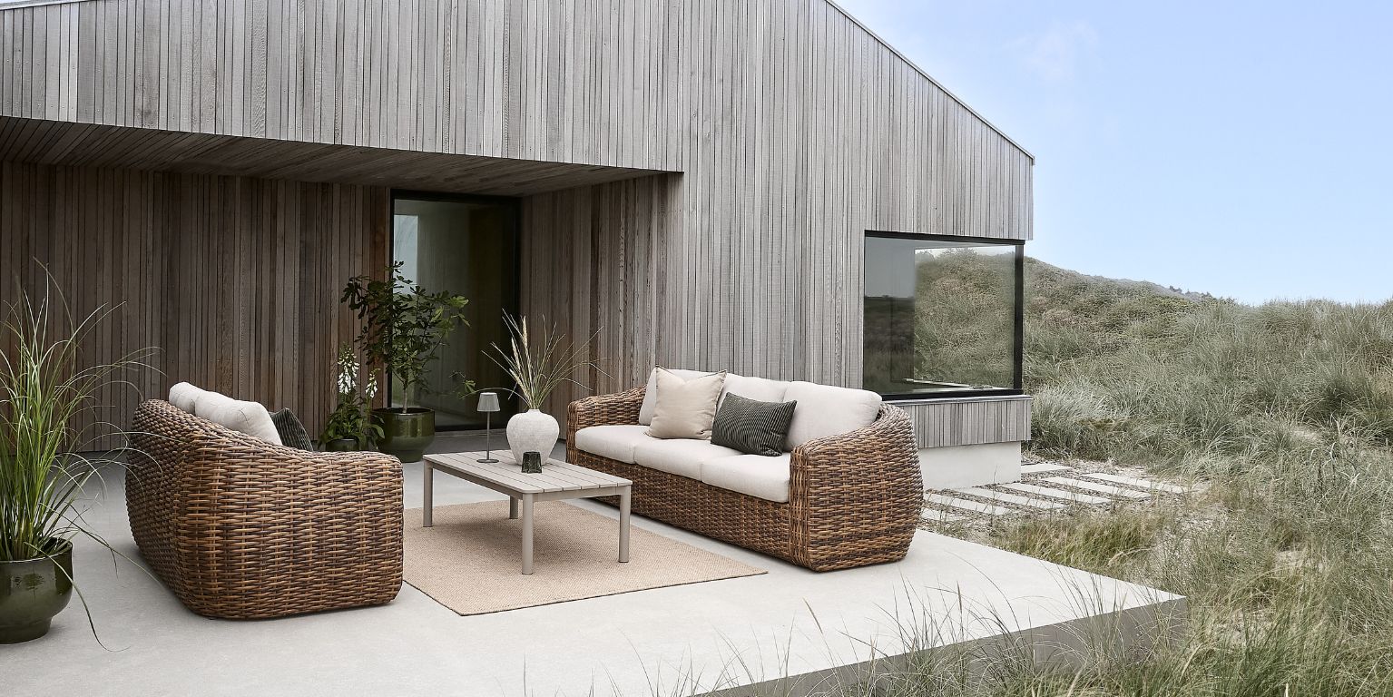 Moderna lounge möbler i flätat material med kuddar placerade på en terrass vid ett modernt hus omgivet av natur.