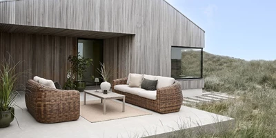 Curva Moderna lounge möbler i flätat material med kuddar placerade på en terrass vid ett modernt hus omgivet av natur.