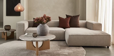 Inredning av ett modernt vardagsrum med en beige modulsoffa, dekorerad med mörkröda och bruna prydkuddar, ett runt soffbord i trä och en stor grå vas.