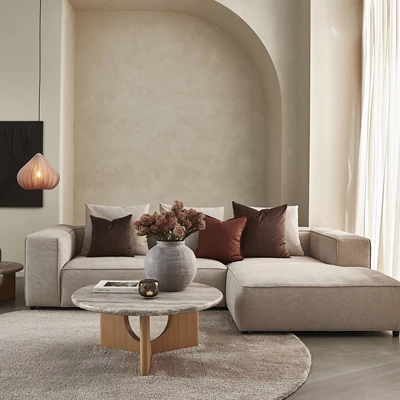 Inredning av ett modernt vardagsrum med en beige modulsoffa, dekorerad med mörkröda och bruna prydkuddar, ett runt soffbord i trä och en stor grå vas.
